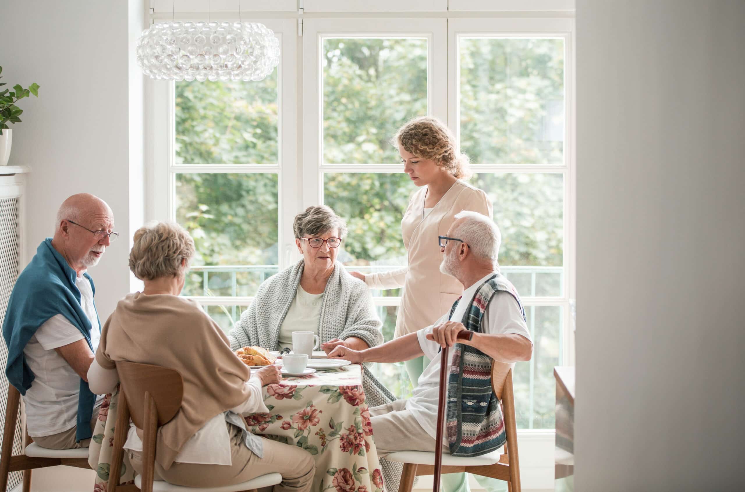 Group of senior friends with helpful carer sitting together at the table at nursing home dining room