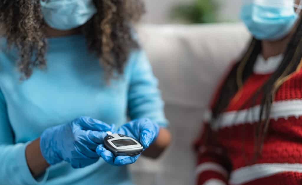 Young nurse checking glycemia test for diabetes to senior patient while wearing surgical gloves and face mask - Healthcare concept